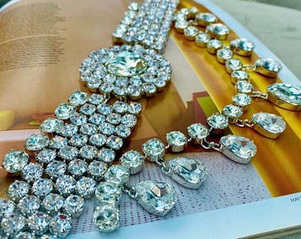 May include: A dazzling display of jewelry featuring a large, ornate necklace with a central oval design and multiple strands of sparkling clear crystals. A second necklace with teardrop-shaped crystals is also visible. The jewelry is set against a magazine page.