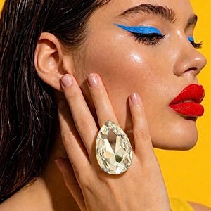 May include: A large, clear, pear-shaped faceted crystal ring worn on the middle finger. The crystal features a pointed top and a rounded base, reflecting light with multiple geometric facets. The ring band is not visible, focusing on the stone.