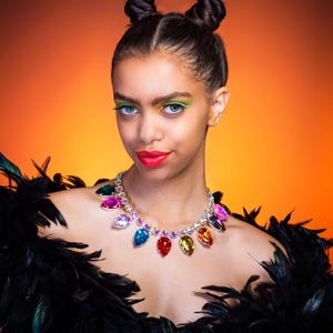 May include: A statement necklace featuring a silver-tone chain adorned with large, multi-coloured, pear-shaped faceted crystals in shades of red, blue, yellow, green, and pink, worn with a black feather garment against a vibrant orange background.
