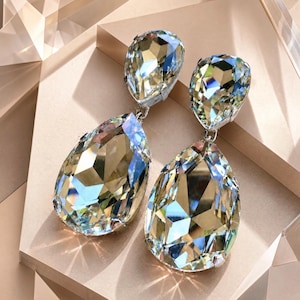 May include: A pair of dazzling, teardrop-shaped earrings. Each earring features a large, faceted crystal pendant in shades of gold, green, and blue, suspended from a smaller, matching crystal top. The earrings are set against a neutral, geometric background.