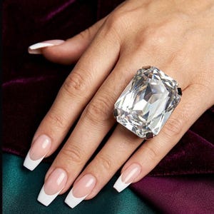 Puede incluir: Un gran anillo rectangular de piedra preciosa transparente con una banda plateada. El anillo está engarzado en una mano con uñas largas y cuidadas con puntas blancas. El fondo es una mezcla de tela morada y verde.