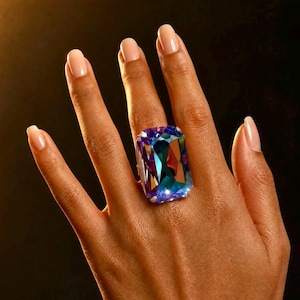 May include: A large rectangular ring featuring a faceted iridescent gemstone that displays shifting hues of deep purple, vibrant blue, and teal. The stone is set in a metallic frame and is positioned on a finger, showcasing its reflective surface.