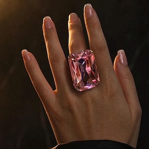 Peut inclure: Une grande bague ornée d'une pierre précieuse rose de taille rectangulaire avec une surface à facettes qui reflète la lumière. La pierre est montée sur un anneau simple, présentant une teinte rose translucide vive, saturée et aux bords nets.
