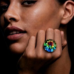 May include: A large, circular ring featuring a faceted, multi-coloured crystal stone. The stone displays a vibrant spectrum of iridescent hues, including shades of blue, green, yellow, and magenta, reflecting light across its polished surface.