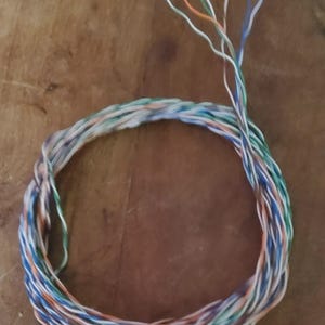 May include: Coiled network cable with multiple colored wires, including blue, orange, green, and white. The wires are twisted together in a circular shape, with some strands extending outwards. The background is a wooden surface.