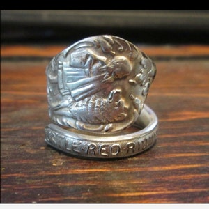 May include: A silver spoon ring with a detailed design of a woman and a wolf. The ring is bent into a circle and the words "Red Riding" are engraved on the band.