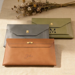 May include: Three leather document cases in shades of brown, grey, and green. Each case features a flap closure with a gold-coloured clasp or lock. The brown case has the name "Julie" inscribed, while the green case has the name "Maximilian".