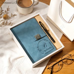 Puede incluir: Un juego de diario y bolígrafo azul en una caja de regalo blanca. El diario tiene un cierre decorativo y el nombre "Belinda" está grabado en la portada. Se incluye un bolígrafo negro con detalles dorados. Una taza de café y gafas están en el fondo.