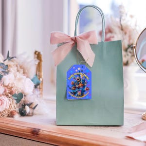 以下が含まれることがあります： ピンクのリボンと「Thank you for coming」と書かれたタグが付いた、薄緑色のギフトバッグ。タグにはキャラクターの漫画の画像があります。バッグは、花と鏡のある窓の近くにあります。