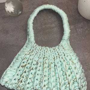 May include: A light blue, handmade crochet bag with a rounded handle. The bag features gold metallic thread woven throughout the crochet, adding a touch of sparkle. The bag's shape flares out from the handle, creating a unique silhouette.