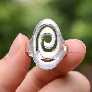 May include: A silver ring with a spiral design. The ring has an oval shape with a cutout spiral in the center. The ring is held in a hand against a blurred green background.