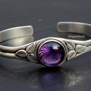 Pode incluir: Uma pulseira de punho prateada com uma grande pedra de ametista roxa escura e redonda. A pulseira tem um design de folhas em ambos os lados da pedra preciosa. O metal tem um acabamento polido. A pulseira é aberta.