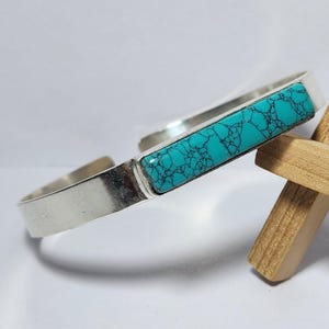 Könnte beinhalten: Ein silbernes Armreif-Armband mit einem rechteckigen Türkisstein. Der Türkisstein hat ein dunkles, netzartiges Muster. Das Armband ist offen und hat ein schlichtes, modernes Design.