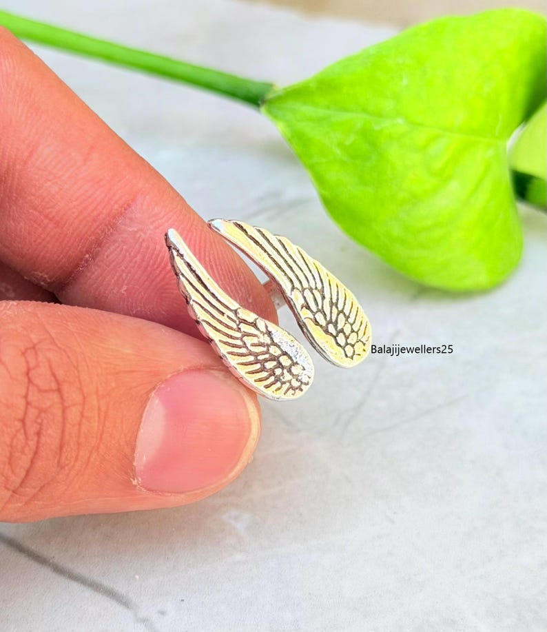 May include: Silver-toned wing-shaped earrings with detailed feather designs. The earrings have a two-tone appearance, with a silver front and a gold-colored back. The earrings are held by a person's finger. The text "Balajijewellers25" is visible.