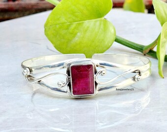 Puede incluir: Una pulsera de plata con un centro de piedra preciosa rectangular de color rojo intenso. La pulsera presenta adornos en espiral y pequeños detalles redondos. La piedra preciosa es el punto focal, engastada en un marco cuadrado.