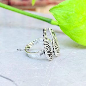 May include: A silver ring featuring a wing design. The ring has two detailed wings that extend upwards. The band of the ring is simple and smooth. The ring is on a light-colored surface with a green leaf in the background.