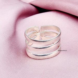 Puede incluir: Un anillo de plata con un diseño multibanda. El anillo presenta tres bandas paralelas conectadas por soportes verticales, creando un aspecto abierto y moderno. El anillo se muestra sobre un fondo de tela rosa.