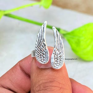 May include: A silver-colored ring featuring an open design with two detailed wing shapes. The wings are textured with feather-like patterns. The ring is held by a person's finger, with a green leafy background.