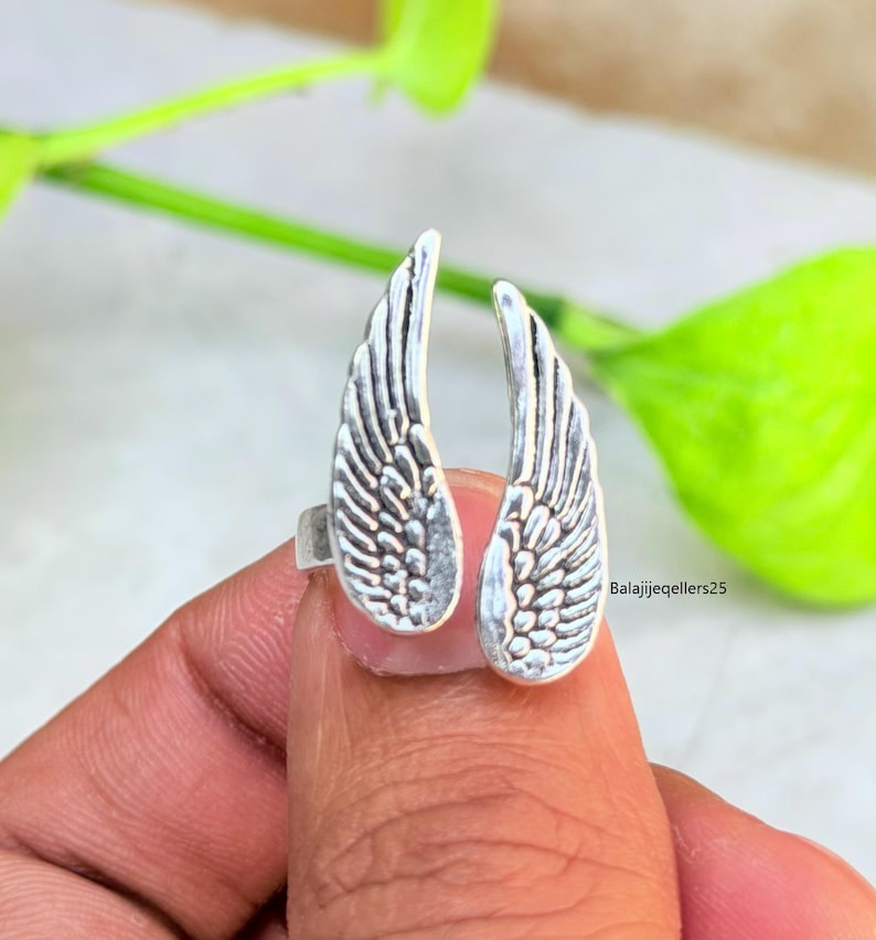 May include: A silver-colored ring featuring an open design with two detailed wing shapes. The wings are textured with feather-like patterns. The ring is held by a person's finger, with a green leafy background.