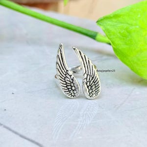 May include: A silver-colored ring featuring a design of two open wings. The wings are detailed with feather patterns. The ring is set against a light-colored surface with green foliage in the background. The text "Balajijeqellers25" is visible.