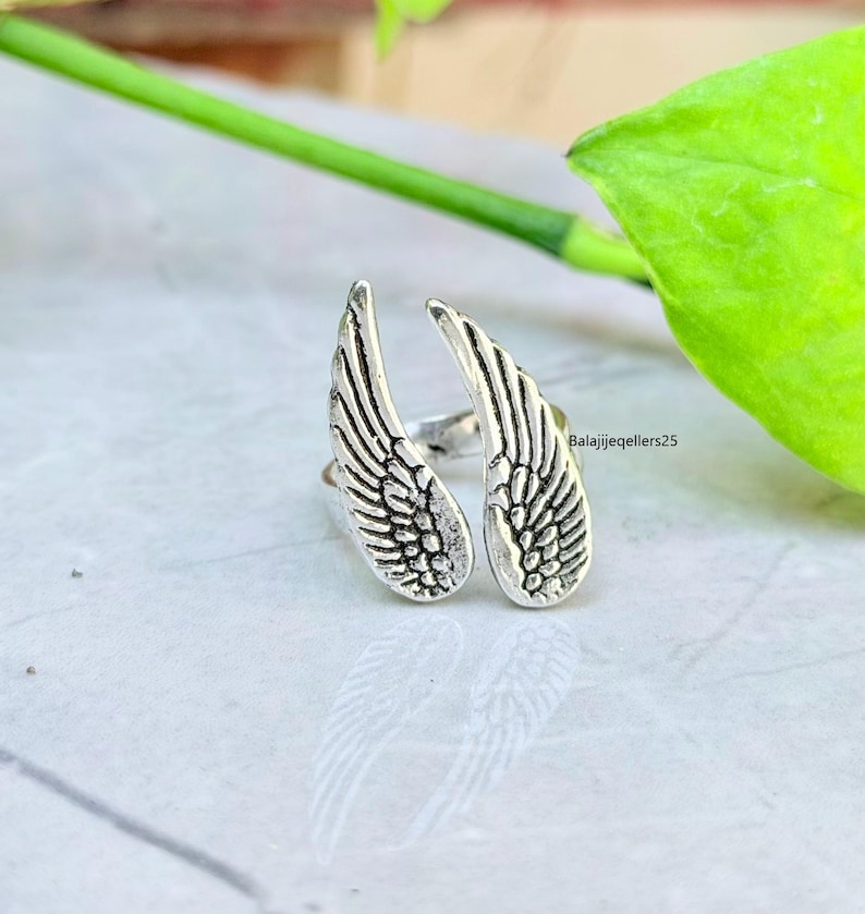 May include: A silver-colored ring featuring a design of two open wings. The wings are detailed with feather patterns. The ring is set against a light-colored surface with green foliage in the background. The text "Balajijeqellers25" is visible.
