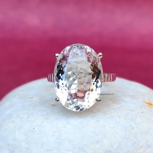 Pode incluir: Anel com uma pedra preciosa transparente de corte oval, montado numa banda prateada. A pedra, facetada, reflete a luz, mostrando a sua clareza. O anel está sobre uma superfície branca, com fundo rosa. Joia.