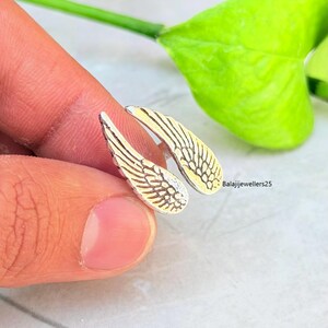 May include: Silver-toned wing-shaped earrings with detailed feather designs. The earrings have a two-tone appearance, with a silver front and a gold-colored back. The earrings are held up by a finger, with a green leaf in the background.