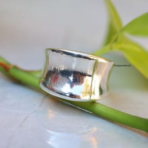 May include: A wide, polished silver ring with a curved design. The ring is set against a blurred background of green leaves and a white surface. The ring's surface reflects the surrounding environment. The text "Balajijewellers25" is visible in the background.