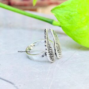 May include: A silver ring featuring a wing design. The ring has two detailed wings that extend upwards. The band of the ring is simple and smooth. The ring is on a light-colored surface with a green leaf in the background.