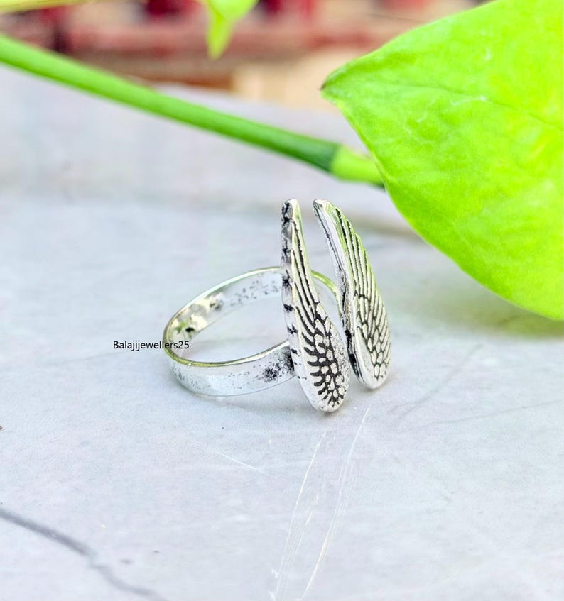 May include: A silver ring featuring a wing design. The ring has two detailed wings that extend upwards. The band of the ring is simple and smooth. The ring is on a light-colored surface with a green leaf in the background.