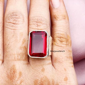 May include: A silver ring featuring a large, rectangular, deep red gemstone. The ring has a thick band and a simple, modern design. The text "Balajijewellers25" is visible on the ring.