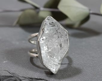May include: A silver ring featuring a large, clear, irregularly shaped crystal. The crystal is set in a silver band with a double-band design. The ring is displayed on a dark gray surface, with a blurred background of green leaves.