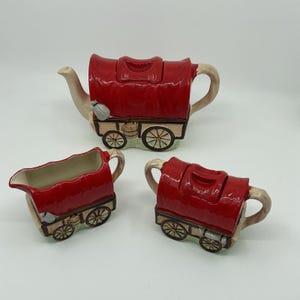 Peut inclure: Un service à thé en céramique composé d'une théière, d'un pot à lait et d'un sucrier, tous en forme de chariots couverts rouges. Chaque pièce présente un dessus rouge brillant, une base beige effet bois, des roues noires et des détails peints.
