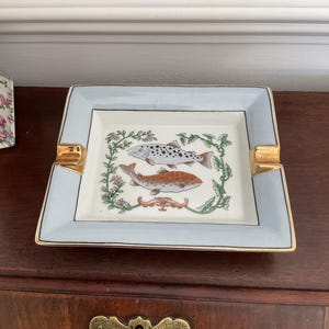 May include: A square ceramic ashtray with a light blue border and gold trim. The center features a white base with a hand-painted illustration of two fish surrounded by green vines and small pink flowers, including two gold-toned cigarette rests.