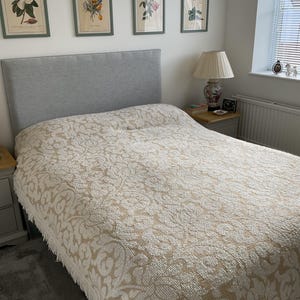 Puede incluir: Una cama con un cabecero tapizado en gris claro y una colcha texturizada en color crema y beige con un patrón floral repetitivo y flecos en los bordes. La cama está situada en una habitación con láminas botánicas enmarcadas en la pared.