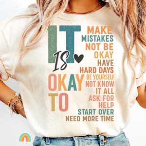 May include: A cream-coloured t-shirt with the words "IT IS OKAY TO" in large, colourful letters. The shirt also features the phrases "MAKE MISTAKES," "NOT BE OKAY," and "HAVE HARD DAYS."