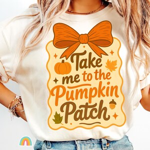 Könnte beinhalten: Cremefarbenes T-Shirt mit einem herbstlichen Motiv. Das Design zeigt eine orangefarbene Schleife, einen Kürbis und den Text "Take me to the Pumpkin Patch" in Braun und Orange. Enthält eine Blatt- und Eichelillustration.