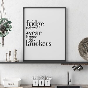 May include: A framed print with the text "Fridge pickers wear bigger knickers" in black font. The print is displayed on a shelf with kitchen accessories, including a hanging plant and utensils.