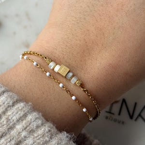 Peut inclure: Deux bracelets dorés sur un poignet. L'un est une chaîne délicate avec de petites perles blanches. L'autre a une chaîne plus épaisse avec une perle carrée dorée avec la lettre "E" et d'autres perles blanches.