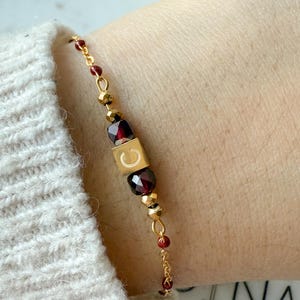 Peut inclure: Bracelet doré orné d'un cube doré avec la lettre "C", de perles rouges facettées et de petites perles dorées. Le bracelet est monté sur une fine chaîne dorée et est présenté sur un poignet.