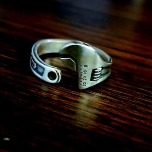 Könnte beinhalten: Ein silberfarbener Ring, der einem Schraubenschlüssel nachempfunden ist. Der Ring weist detaillierte Markierungen und Messungen auf, einschließlich der Zahlen 30, 20, 10 und 0. Der Ring ist auf einer dunklen Holzoberfläche platziert.