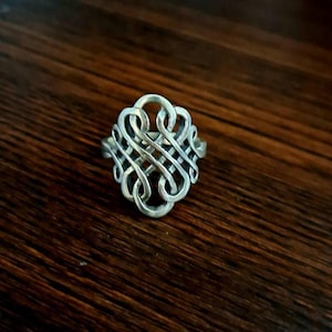 Puede incluir: Un anillo plateado con un diseño intrincado e entrelazado. El anillo presenta una serie de formas entrelazadas, creando un patrón complejo. El anillo se encuentra sobre una superficie de madera oscura, resaltando su brillo metálico.
