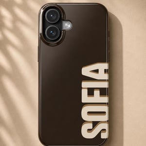 May include: A dark brown smartphone case featuring the name SOFIA printed in large, vertical, cream-colored block letters with a brown shadow effect. The case has a cutout for a dual-camera system and a circular flash on the upper left side.