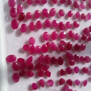 May include: A collection of numerous small, loose, faceted rubies in various oval and pear shapes. The gemstones exhibit a vibrant, deep pinkish-red hue and are arranged in rows on a white display tray with a raised border, showcasing their clarity.