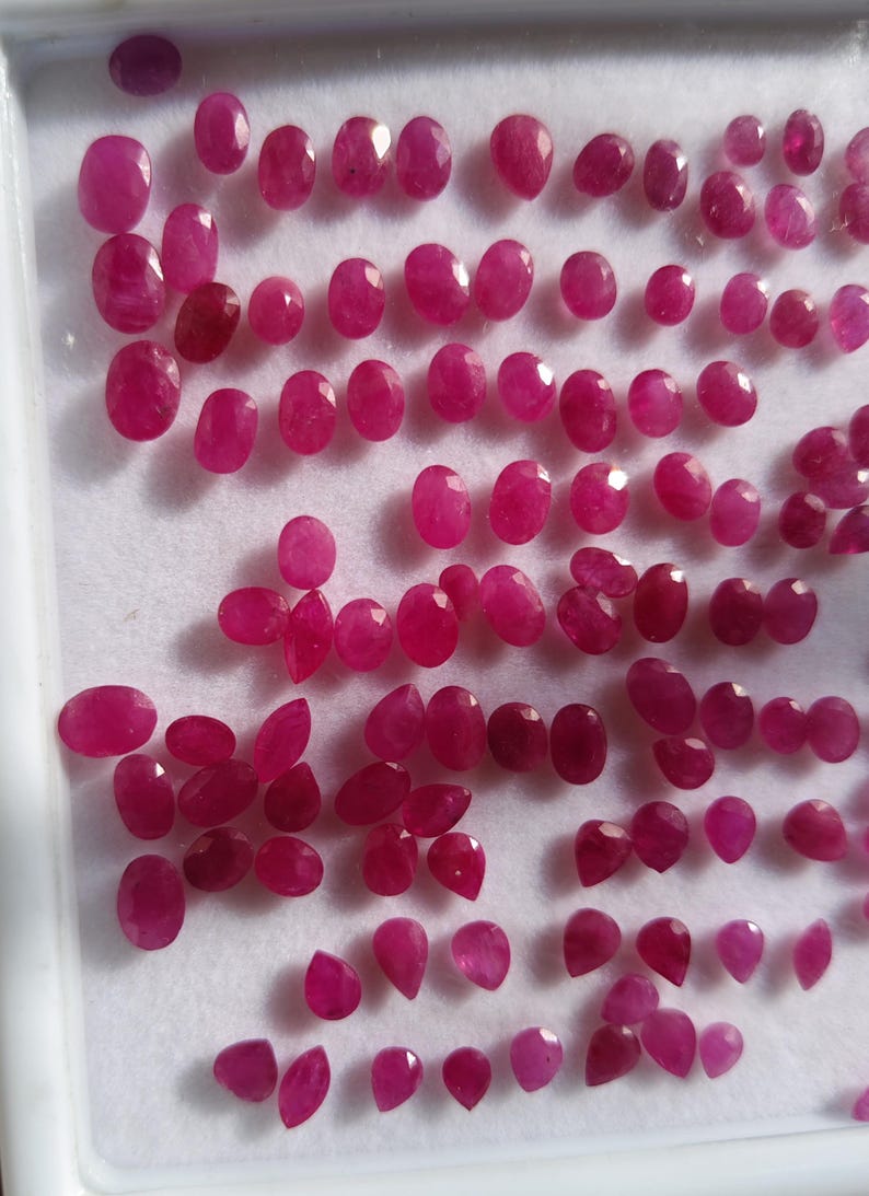 May include: A collection of numerous small, loose, faceted rubies in various oval and pear shapes. The gemstones exhibit a vibrant, deep pinkish-red hue and are arranged in rows on a white display tray with a raised border, showcasing their clarity.