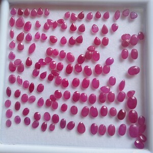 May include: A collection of numerous small, faceted, vibrant pinkish-red gemstones arranged in a white display tray. The stones vary in shape, including oval, pear, and round cuts, all exhibiting a polished, reflective surface and consistent colour.