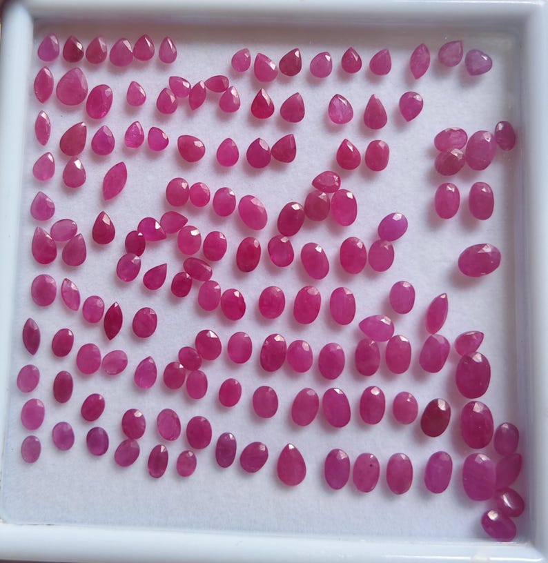 May include: A collection of numerous small, faceted, vibrant pinkish-red gemstones arranged in a white display tray. The stones vary in shape, including oval, pear, and round cuts, all exhibiting a polished, reflective surface and consistent colour.