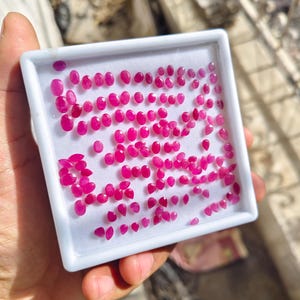May include: A square white display box containing a large collection of small, faceted, oval and pear-shaped loose gemstones in a vibrant magenta-pink colour. The stones are arranged in rows on a white foam insert within the plastic container.