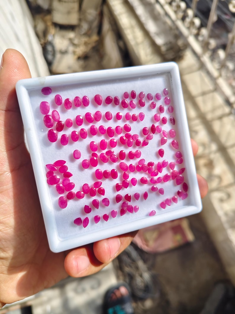 May include: A square white display box containing a large collection of small, faceted, oval and pear-shaped loose gemstones in a vibrant magenta-pink colour. The stones are arranged in rows on a white foam insert within the plastic container.