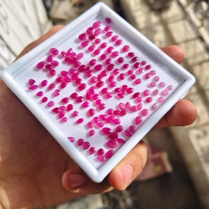 May include: A white square display tray containing a large collection of small, faceted, vibrant pink and red gemstones. The stones are arranged in a loose cluster on a white foam insert, featuring various shapes including oval, pear, and round cuts.